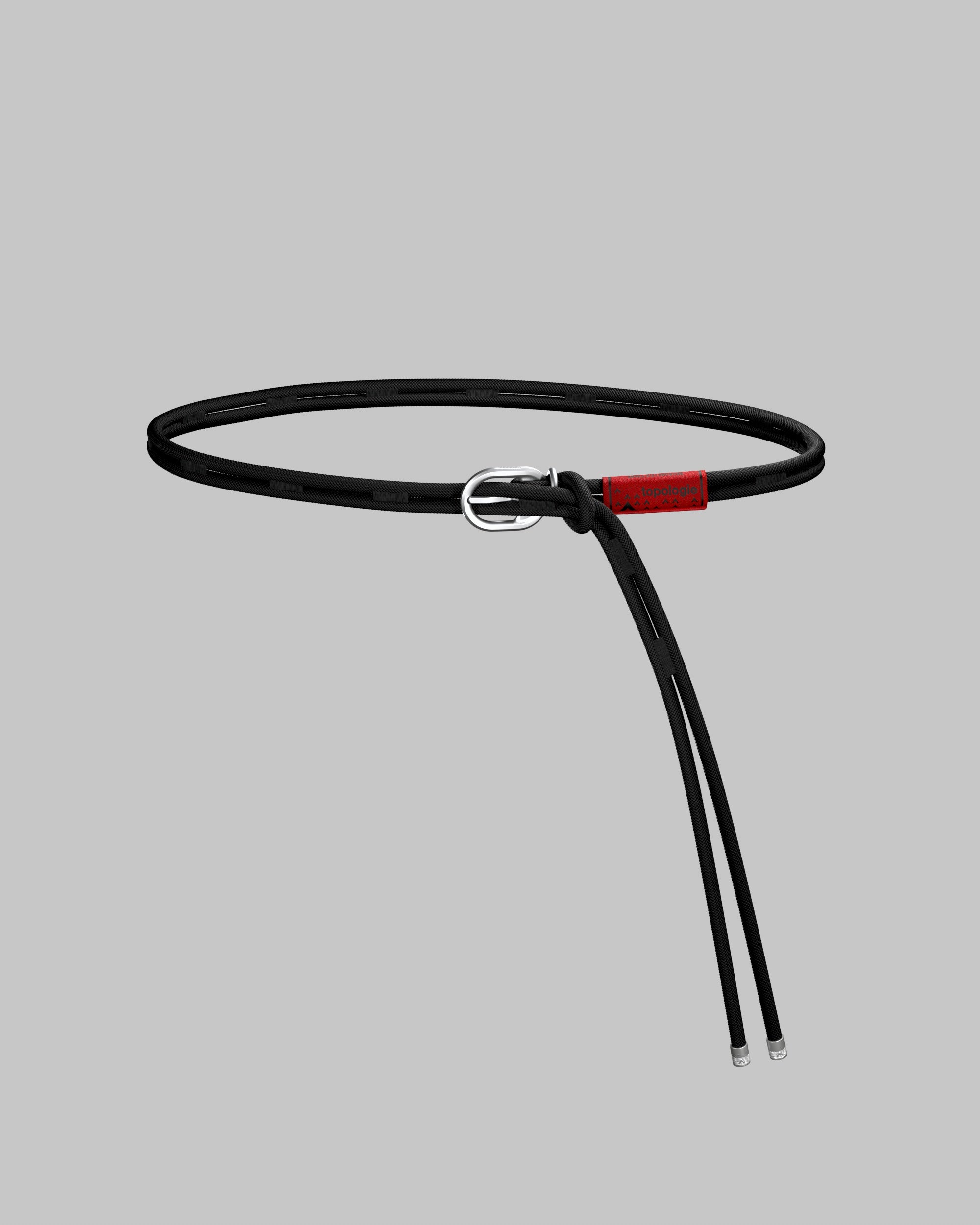 Knot Belt in thin black cord with a silver metal buckle, red topologie tag, and silver cord tips, shown against a light grey background.

