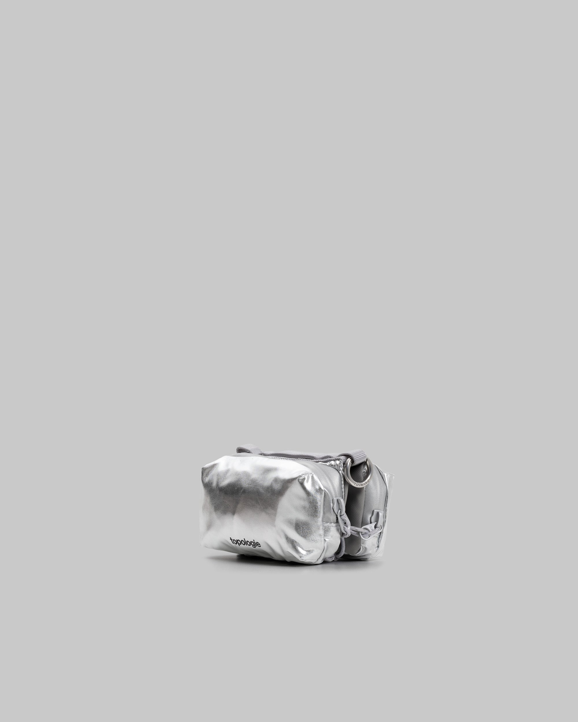 Mini Bottle Sacoche in metallic silver with a black topologie logo and silver D-ring, shown against a light grey background.