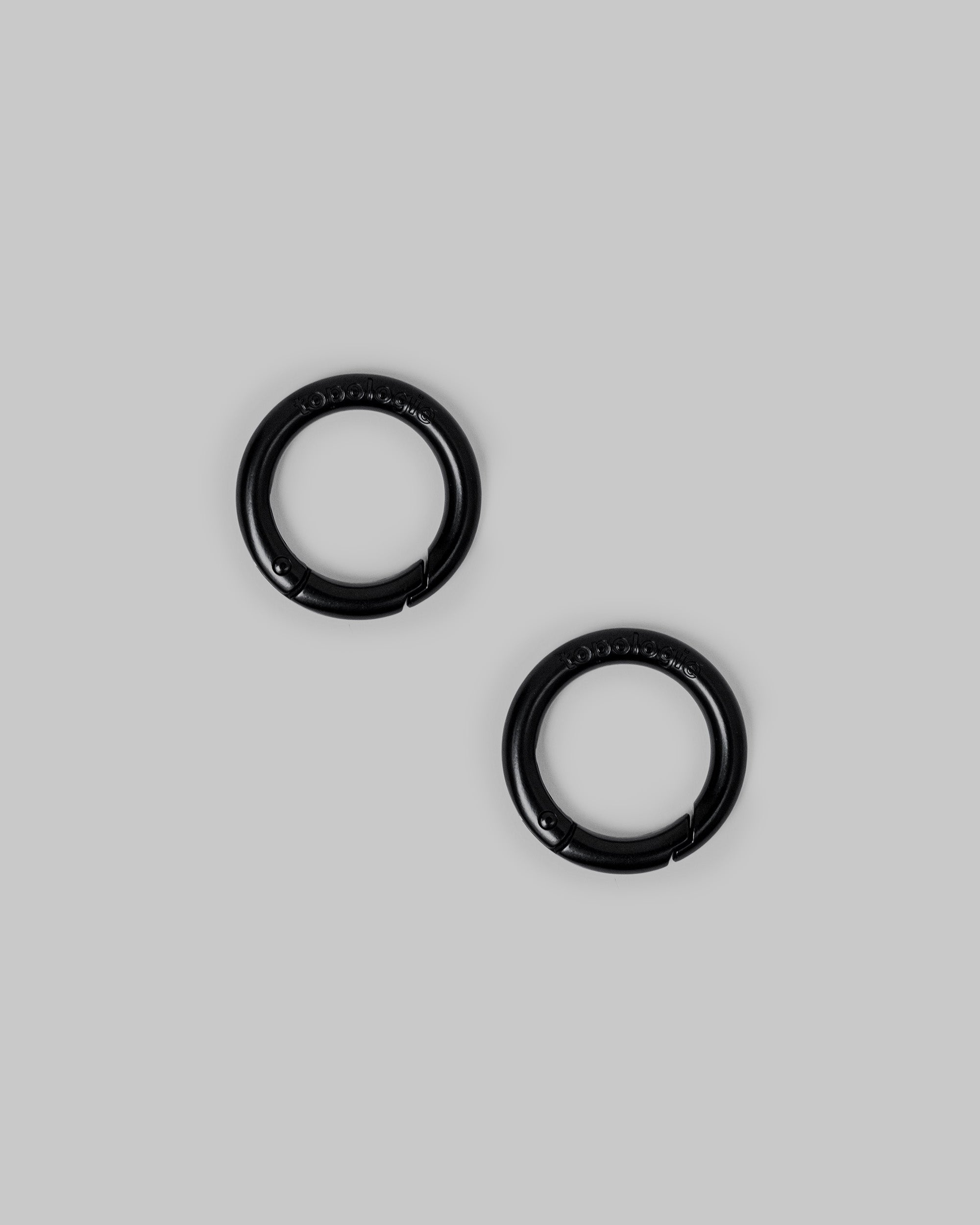 O-Ring in matte black metal with embossed topologie logos and spring-gated closures, shown as a pair against a light grey background.