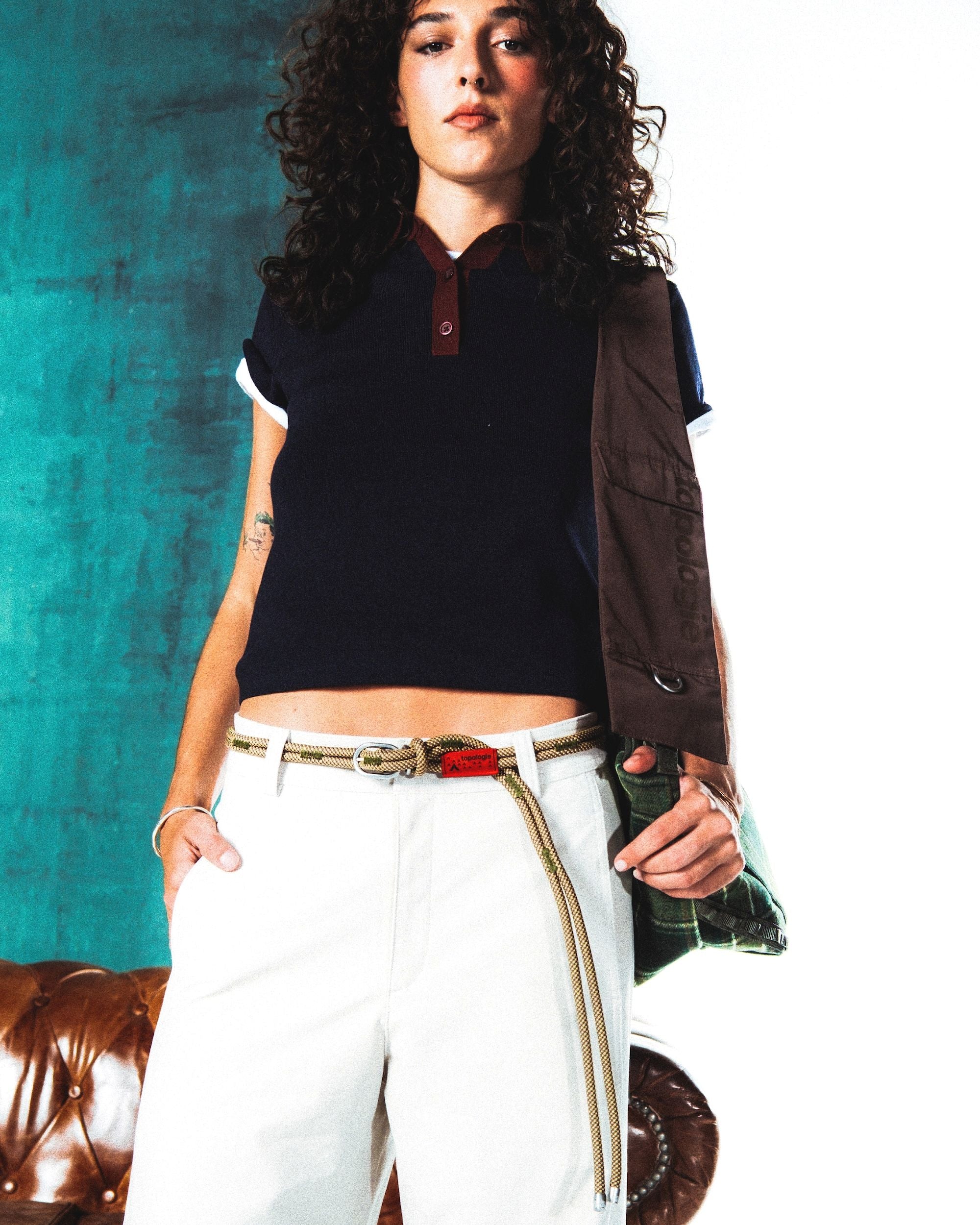 A person wearing a navy short-sleeve polo shirt, white pants, and a rope belt with a red tag, carrying a brown and green bag over their shoulder.
